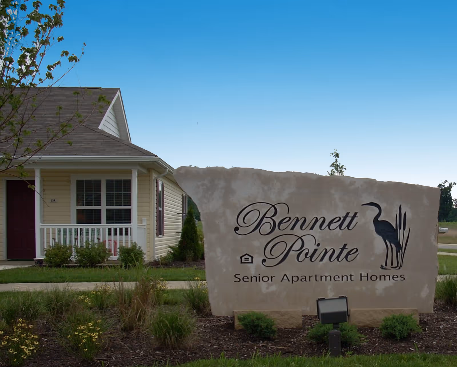 Stone sign for Bennett Pointe Senior Apartment Homes with a silhouette of a bird and reeds, situated in front of a beige house with a porch and a maroon door under a clear blue sky.