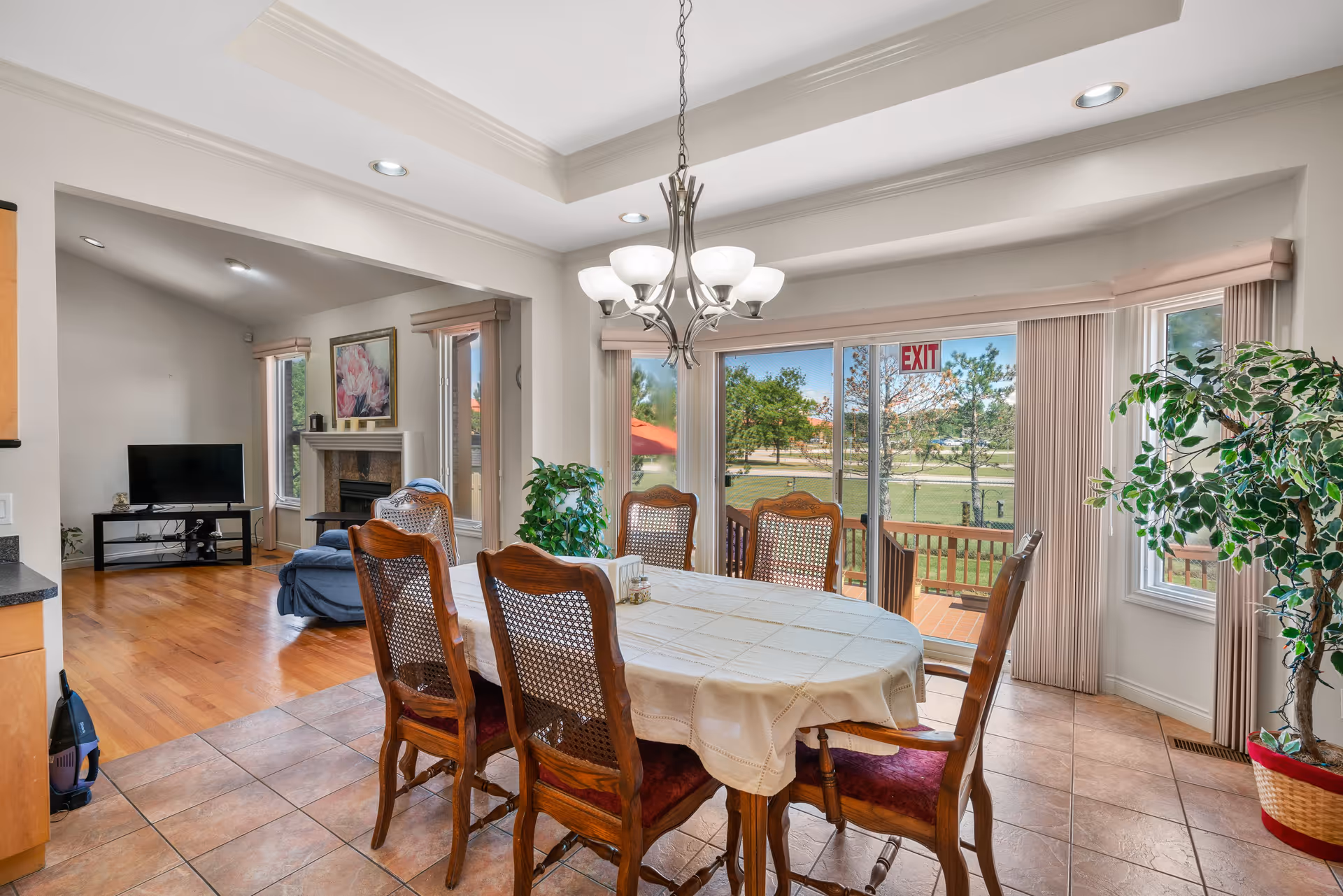 A dining area with a wooden table covered by a white tablecloth and six wooden chairs with red cushions. A chandelier with multiple lights hangs above the table. Large sliding glass doors with vertical blinds lead to an outdoor patio with a view of green grass and trees. To the left, there is a living room area with a TV on a stand, a fireplace, and a blue armchair. The floor is tiled in the dining area and hardwood in the living room. A potted plant is near the window on the right.