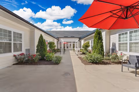 Outdoor courtyard area of Morningside House of Fredericksburg with a clear blue sky and scattered clouds. The courtyard is surrounded by single-story white buildings with large windows. There are landscaped garden beds with green shrubs and red flowers, and a large red patio umbrella providing shade over a seating area with black chairs.