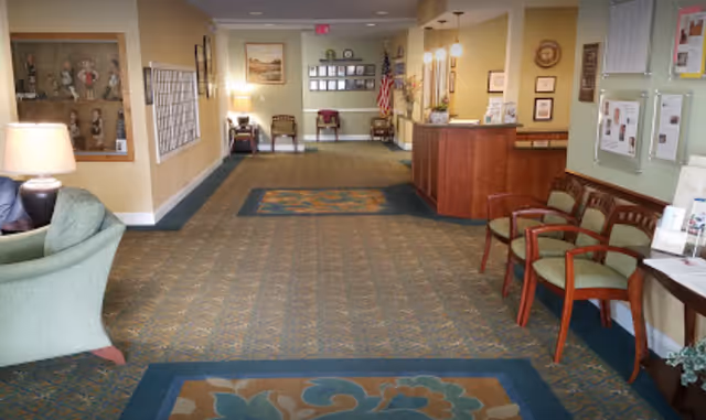 Interior view of a retirement village common area with patterned carpet, green upholstered chairs, wooden reception desk, wall-mounted display cases, framed pictures, and an American flag in the background.