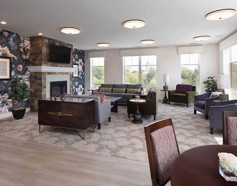 Bright communal lounge with sofas and armchairs arranged around a fireplace and large windows overlooking trees.
