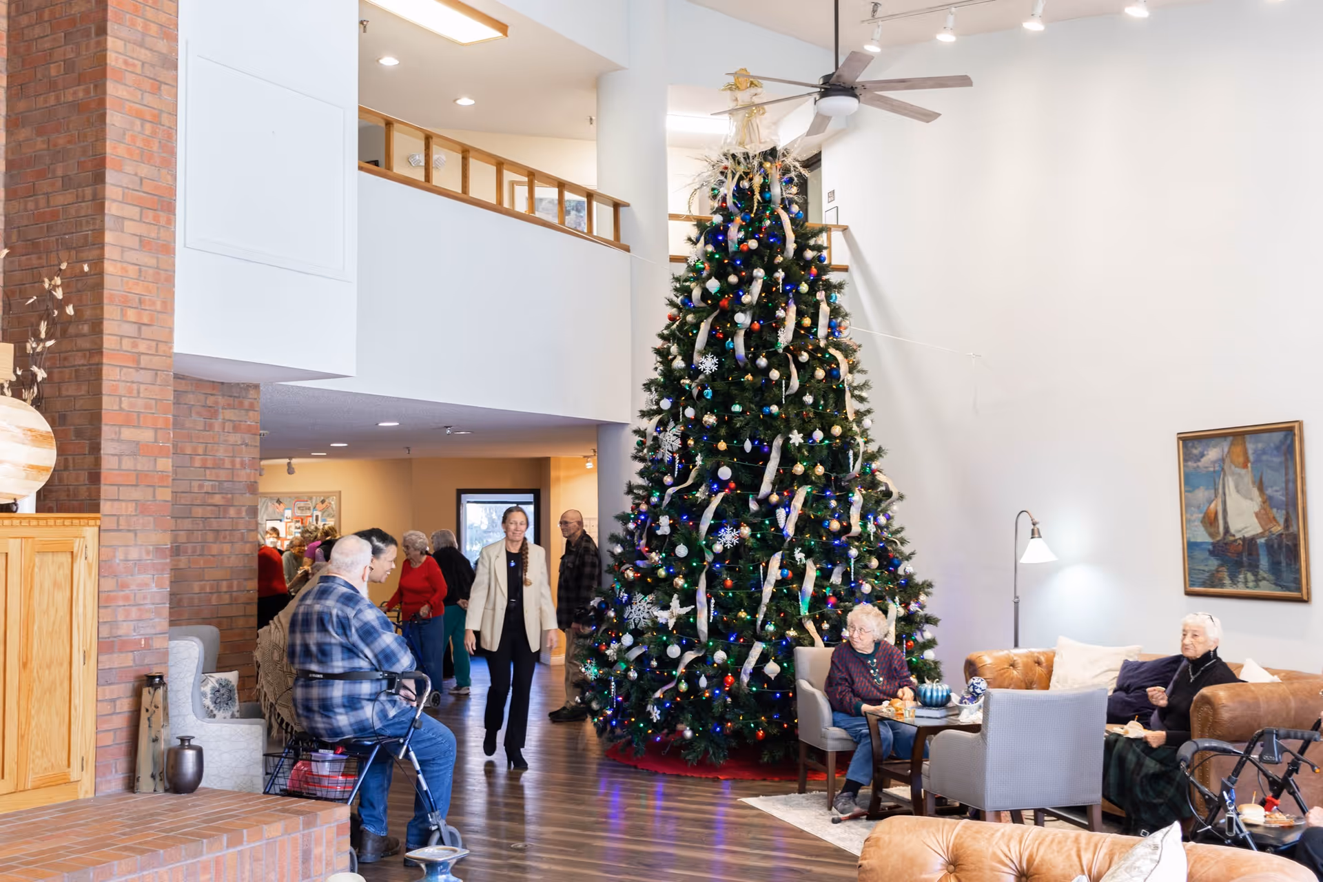 A spacious living room area decorated for Christmas with a large, decorated Christmas tree in the center. Several elderly people are seated on chairs and sofas around the tree, some using walkers. A woman in a beige jacket is walking through the room. The room has wooden flooring, a brick fireplace, and a painting of a sailboat on the wall.