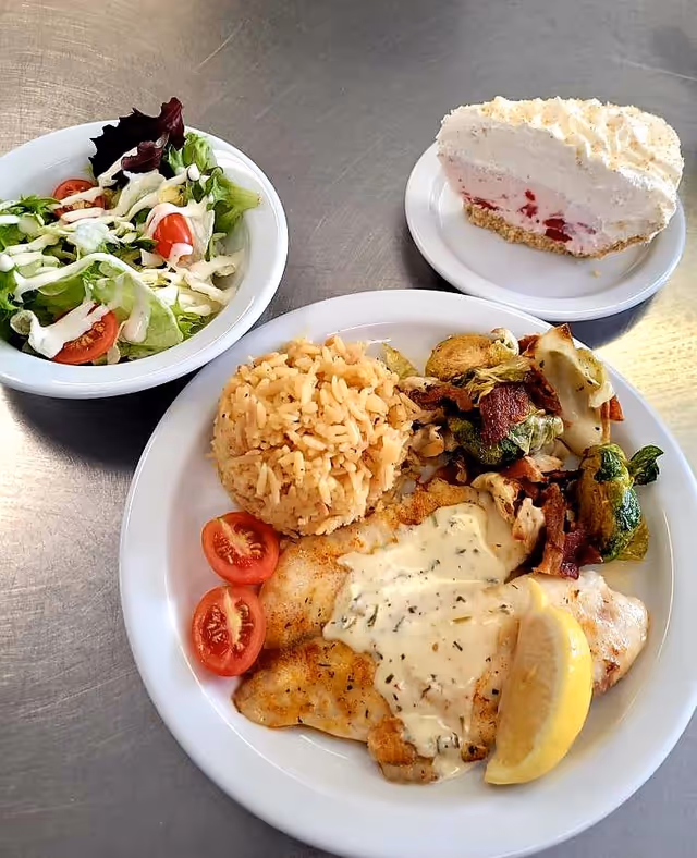 A meal consisting of a plate with grilled fish topped with a creamy herb sauce, a lemon wedge, two cherry tomato halves, a serving of rice, and roasted Brussels sprouts. Next to it is a bowl of fresh salad with lettuce, cherry tomatoes, and dressing. A slice of creamy dessert with a crumbly crust is on a separate small plate.