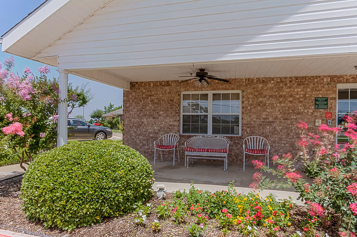 Covered outdoor patio area with a ceiling fan, three white chairs with red cushions, and a small garden with green bushes and colorful flowers in front. A brick wall with a window is visible behind the chairs, and a parked car is seen to the left.
