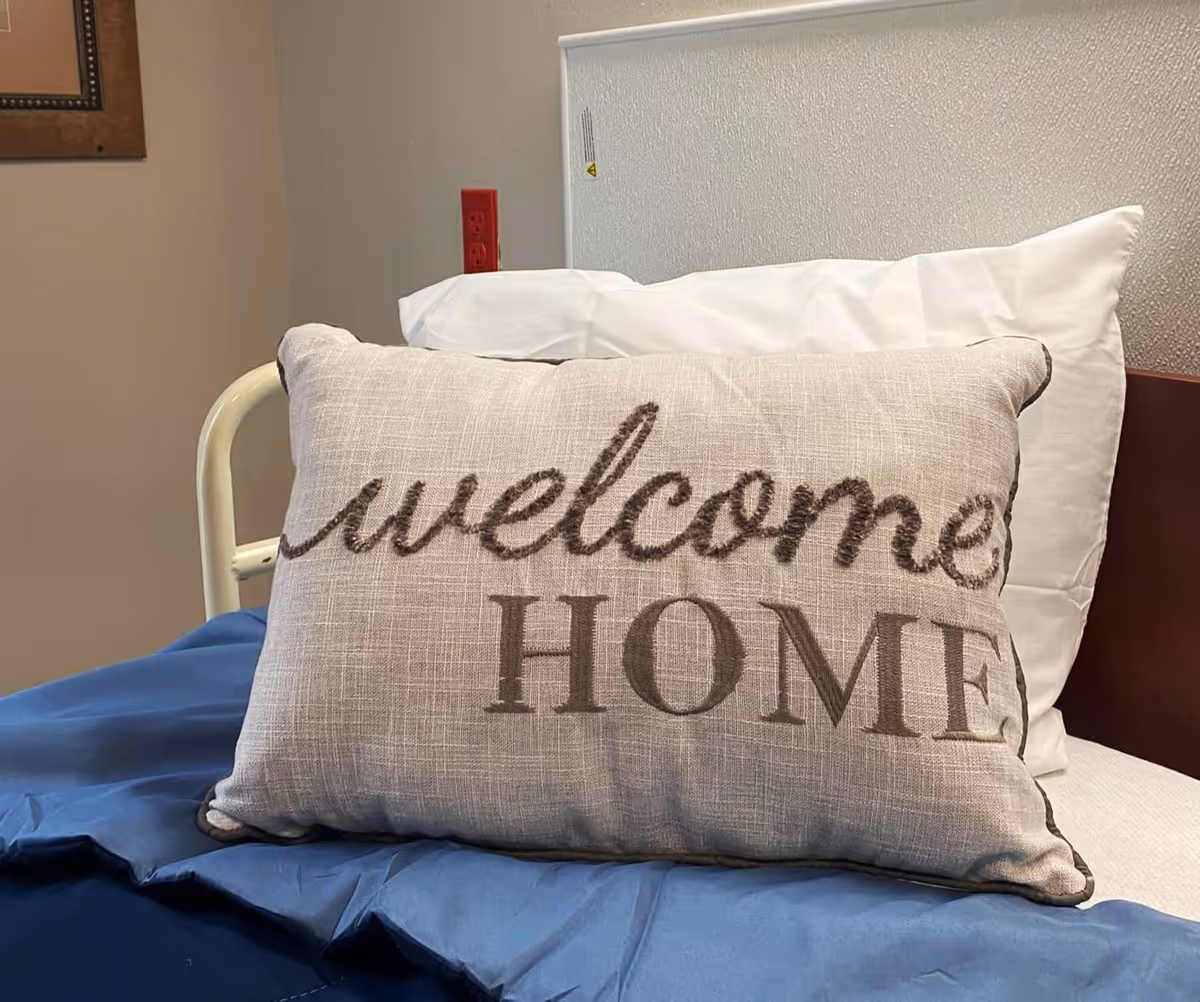A decorative pillow embroidered with "welcome HOME" rests on a bed with white pillows and blue bedding in a room.