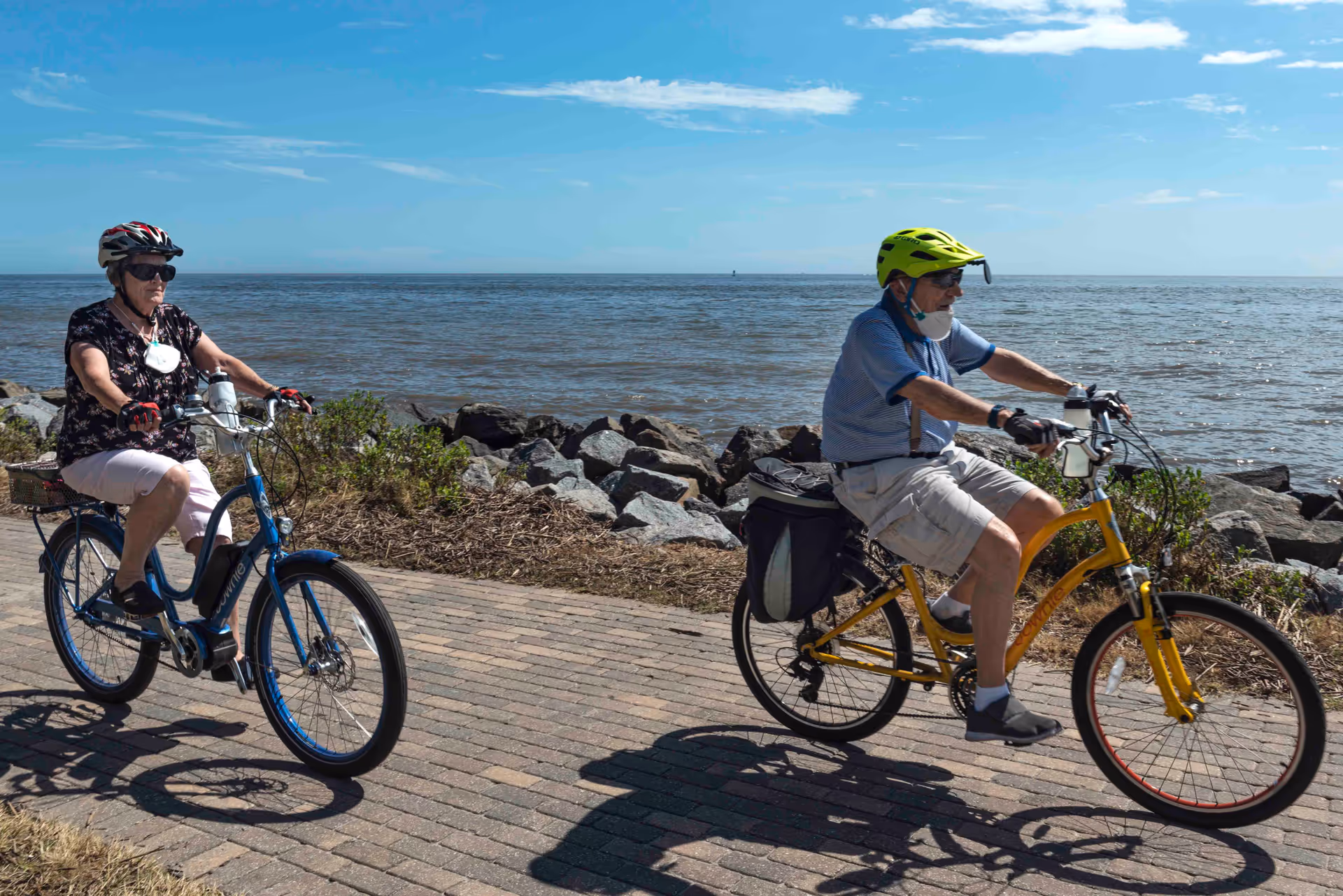 Two elderly people riding bicycles on a paved path along a rocky shoreline with the ocean and blue sky in the background. Both riders are wearing helmets and casual clothing.
