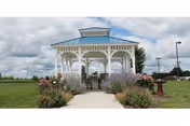 White gazebo at the end of a paved walkway surrounded by flower beds on a grassy lawn under a cloudy sky.