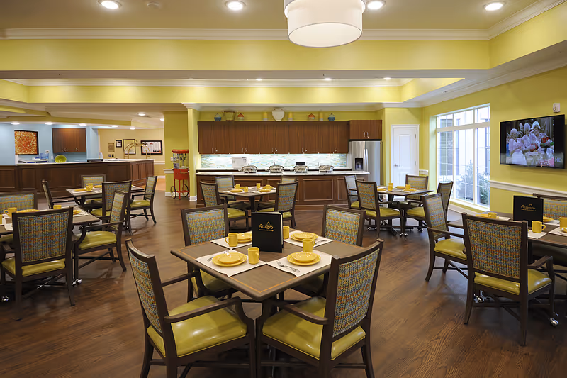 A bright and spacious dining room in a senior living facility with multiple tables set with yellow plates, cups, and utensils. The room features wooden flooring, yellow walls, and large windows letting in natural light. There is a kitchen area with cabinets, a refrigerator, and a buffet setup in the background. A television is mounted on the wall showing a program.