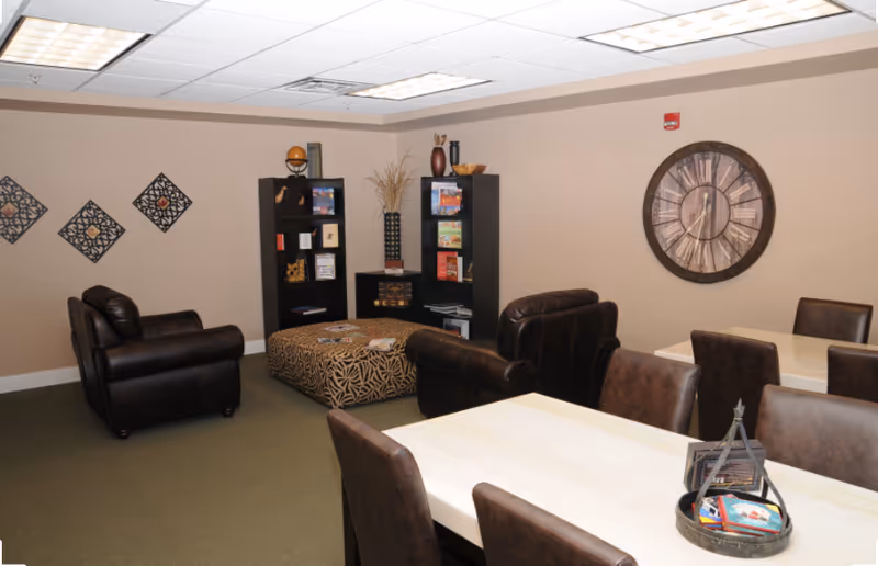 A senior living facility lounge with brown leather seating, tables, a bookshelf, and a decorative clock.