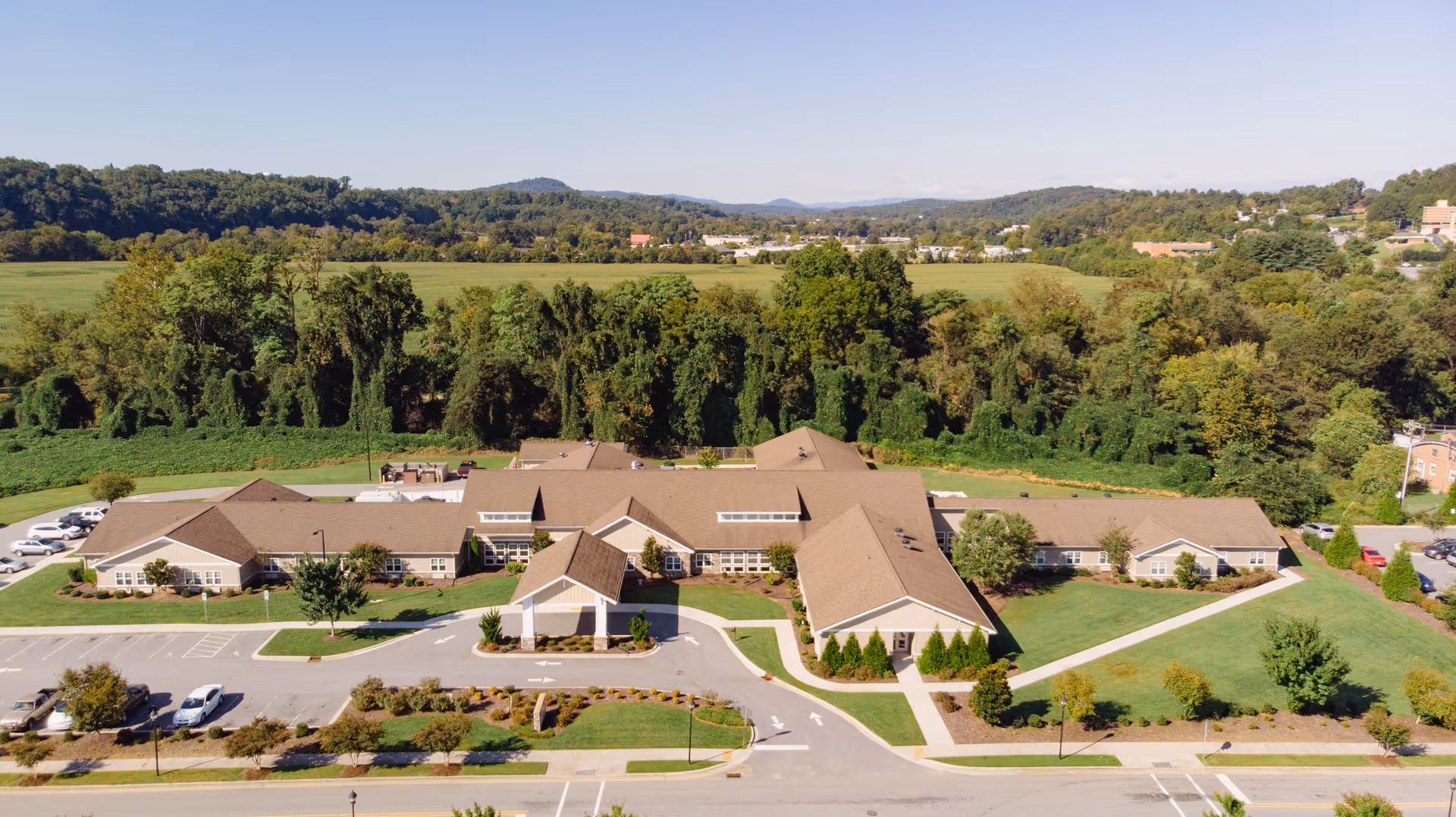 Aerial view of a one-story senior living facility with a porte-cochere, circular drive, parking lot, and landscaped grounds surrounded by trees and fields.