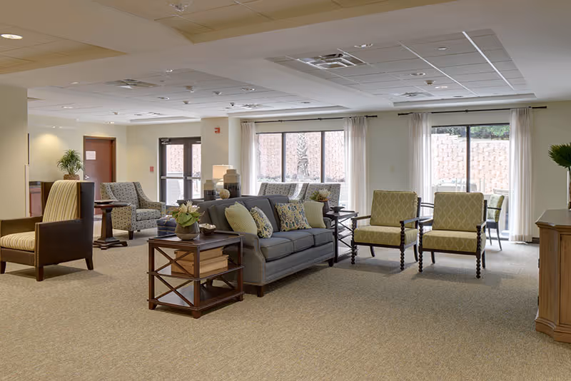 A spacious senior living common area with a gray sofa adorned with patterned cushions, two green armchairs, and additional seating in the background. Large windows with white curtains allow natural light to brighten the room. The space is carpeted and decorated with side tables, plants, and lamps.