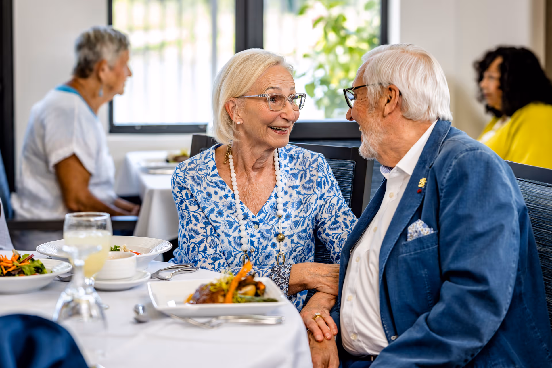 An elderly couple sitting at a dining table in a senior living facility, smiling and holding hands while engaging in conversation. Plates of food and drinks are on the table. Other seniors are visible in the background, seated at tables near windows with natural light.