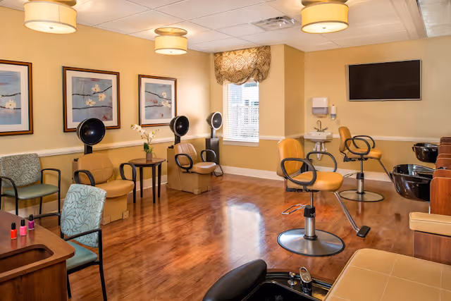 Interior view of a salon area in a rehabilitation center with salon chairs, hair dryers, a manicure station with nail polish, framed artwork on the walls, a window with a patterned valance, and a wall-mounted TV.