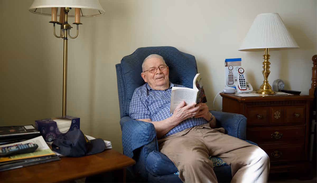 An elderly man wearing glasses and a blue checkered shirt is sitting comfortably in a blue armchair, reading a book and smiling. The room has a wooden side table with a tissue box, a cap, and some magazines, and a wooden dresser with two remote controls and a brass lamp with a white lampshade.
