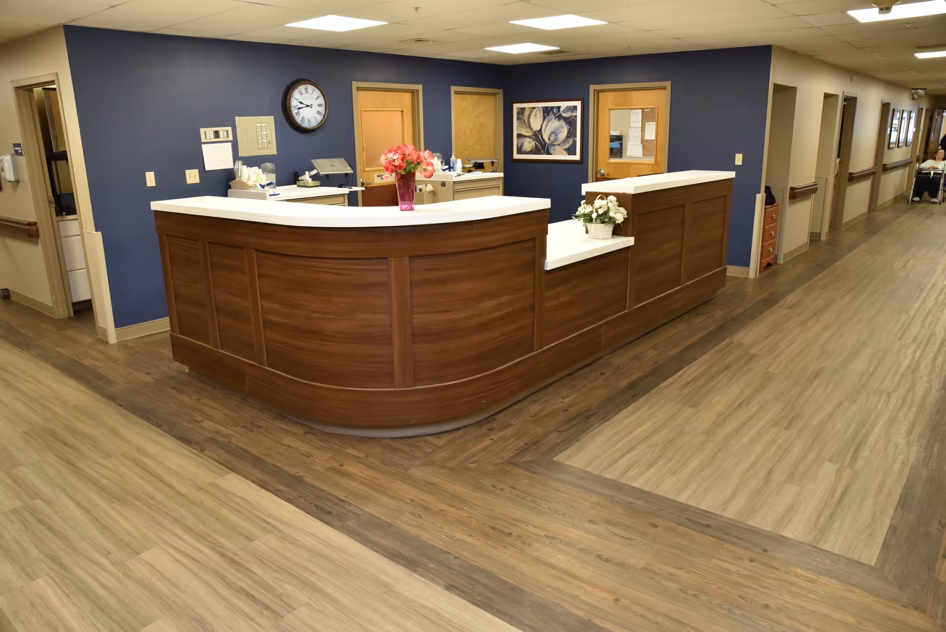 A curved wooden reception/nurses' station in a senior living facility hallway with wood-look flooring and a blue accent wall.