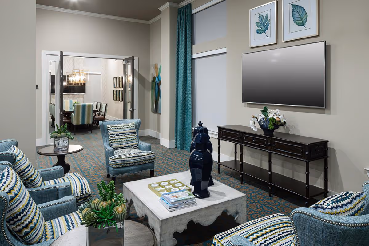 A cozy living room area with patterned blue armchairs and a matching ottoman around a white coffee table. The coffee table has decorative items including a blue vase and a tic-tac-toe game. A dark wooden console table with a flower arrangement sits beneath a mounted flat-screen TV. The room has beige walls, teal curtains, and carpet with a blue and orange pattern. Through open double doors, a dining room with a chandelier and chairs is visible.