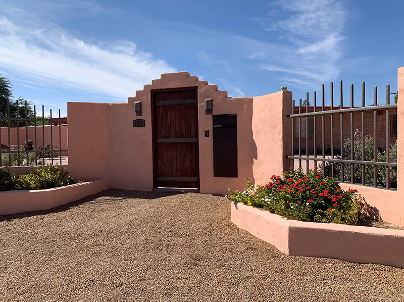 Outdoor view of a gated entrance with a wooden door set in a pink stucco wall. The ground is covered with gravel, and there are raised flower beds with red and white flowers on either side of the entrance. The sky is clear with some clouds.