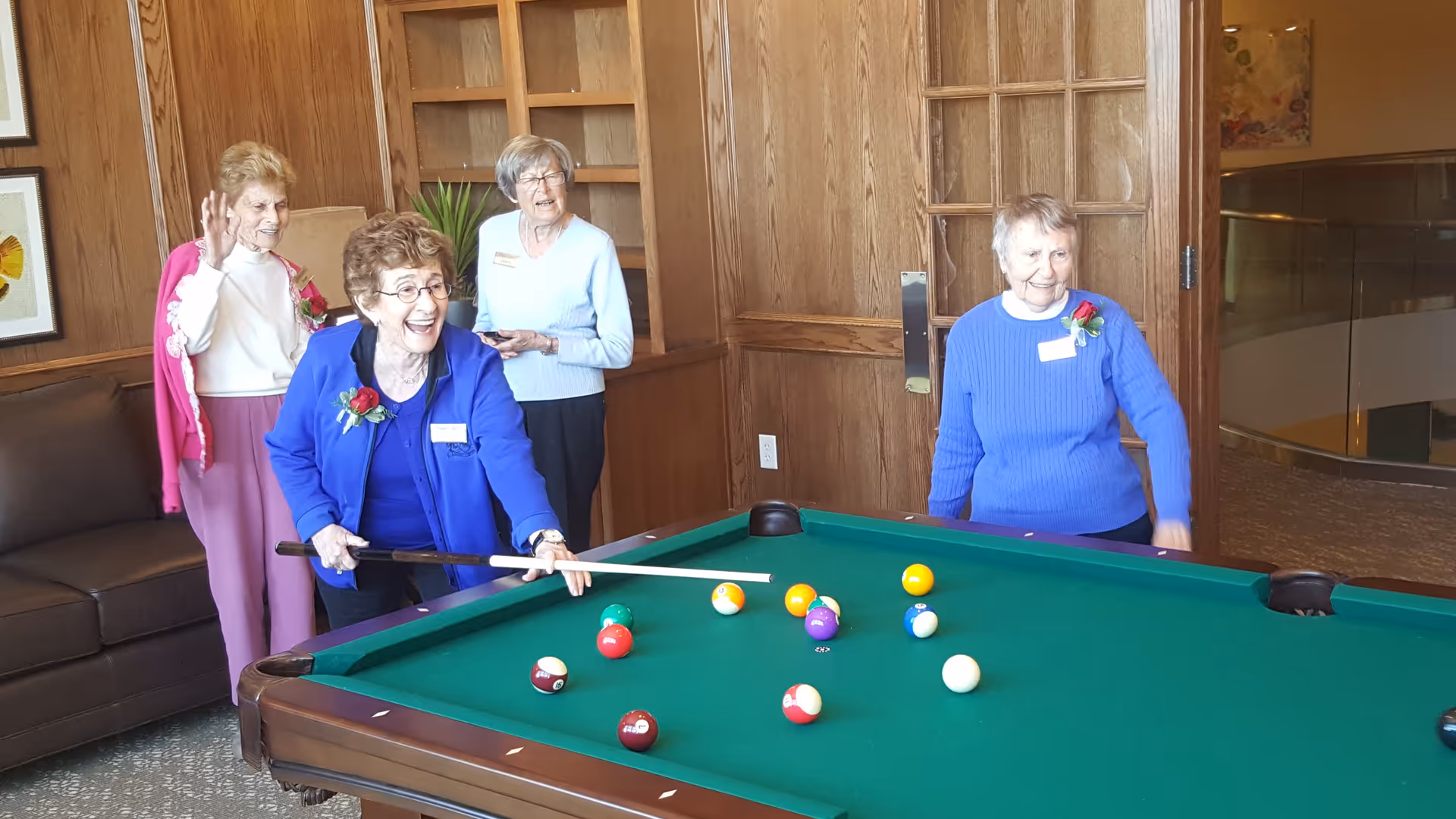 Four elderly women playing pool in a room with wooden panel walls. One woman is holding a pool cue and smiling as she prepares to take a shot, while the others watch and smile. There is a brown couch and framed artwork on the wall behind them.