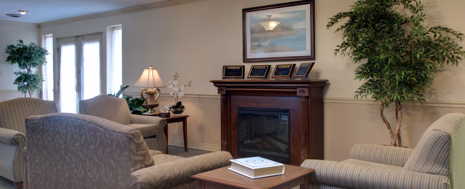 A cozy living room area in a nursing and rehabilitation facility featuring several upholstered armchairs arranged around a wooden coffee table with a book on it. There is a wooden fireplace with plaques on the mantel and a framed picture above it. The room is decorated with potted plants and a table lamp, with natural light coming through glass doors in the background.