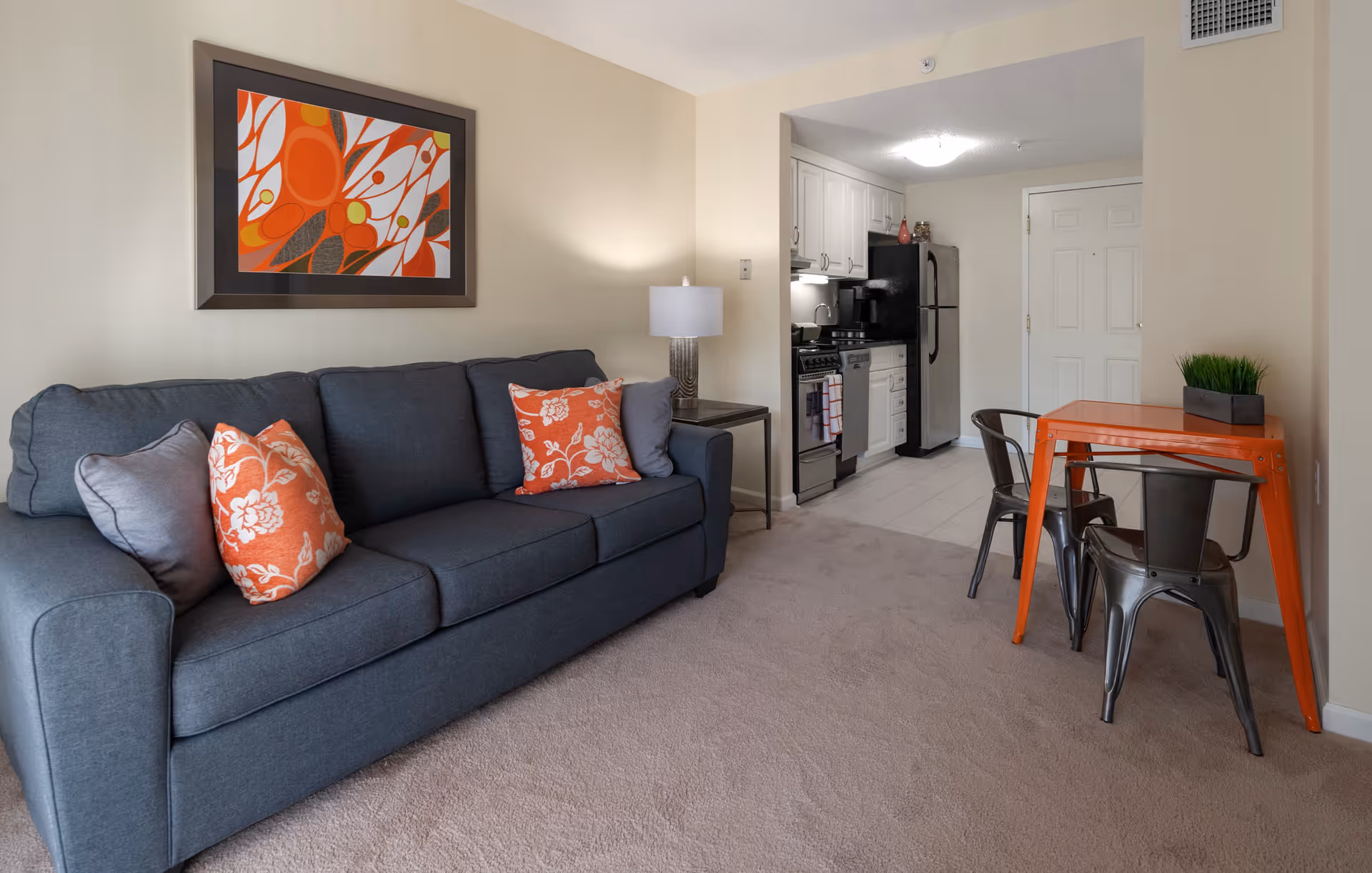 A cozy living area featuring a dark gray sofa with orange and gray floral pillows, a side table with a lamp, and a bright orange dining table with two metal chairs. The background shows a small kitchen with white cabinets, a black refrigerator, and a stove. The walls are light beige and there is a colorful abstract painting above the sofa.