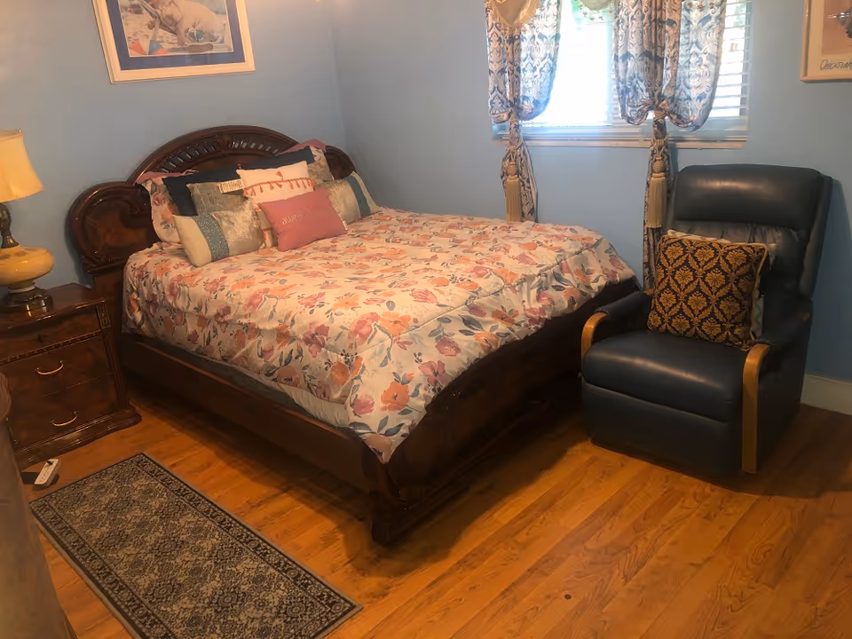A cozy bedroom with a wooden bed frame and floral patterned bedding, multiple decorative pillows, a wooden nightstand with a lamp, a blue recliner chair with a patterned cushion, a window with patterned curtains, and hardwood flooring with a small rug.