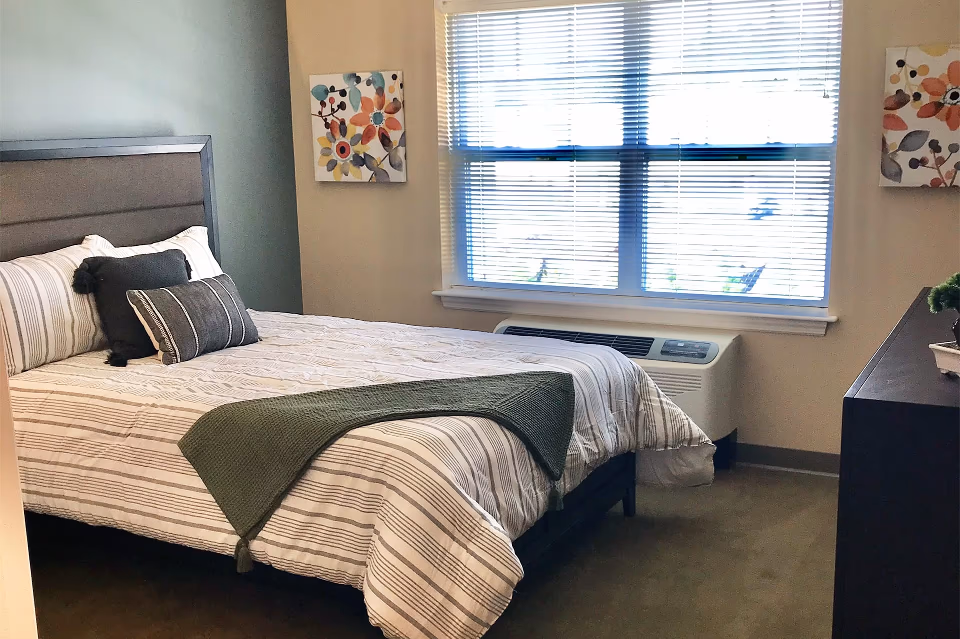 A neatly made bed with striped bedding and two decorative pillows in a bedroom. The room has a large window with blinds, two floral paintings on the walls, a dark wooden dresser with a small plant on top, and a wall-mounted air conditioning unit below the window.