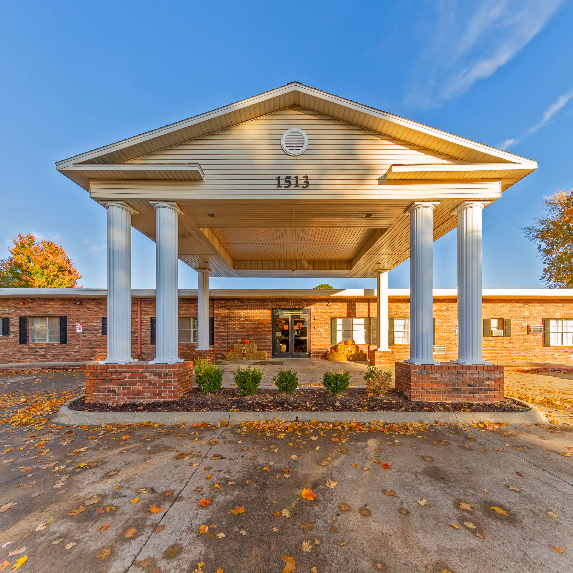 Front exterior view of a single-story brick building with a covered entrance supported by four white columns. The building number 1513 is displayed on the front of the entrance roof. There are small bushes and autumn leaves scattered on the ground in front of the building under a clear blue sky.