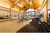 A spacious fitness room with large windows letting in natural light, featuring treadmills, weight machines, free weights, and mirrors along one wall. The room has a high wooden ceiling with exposed beams and ceiling fans.