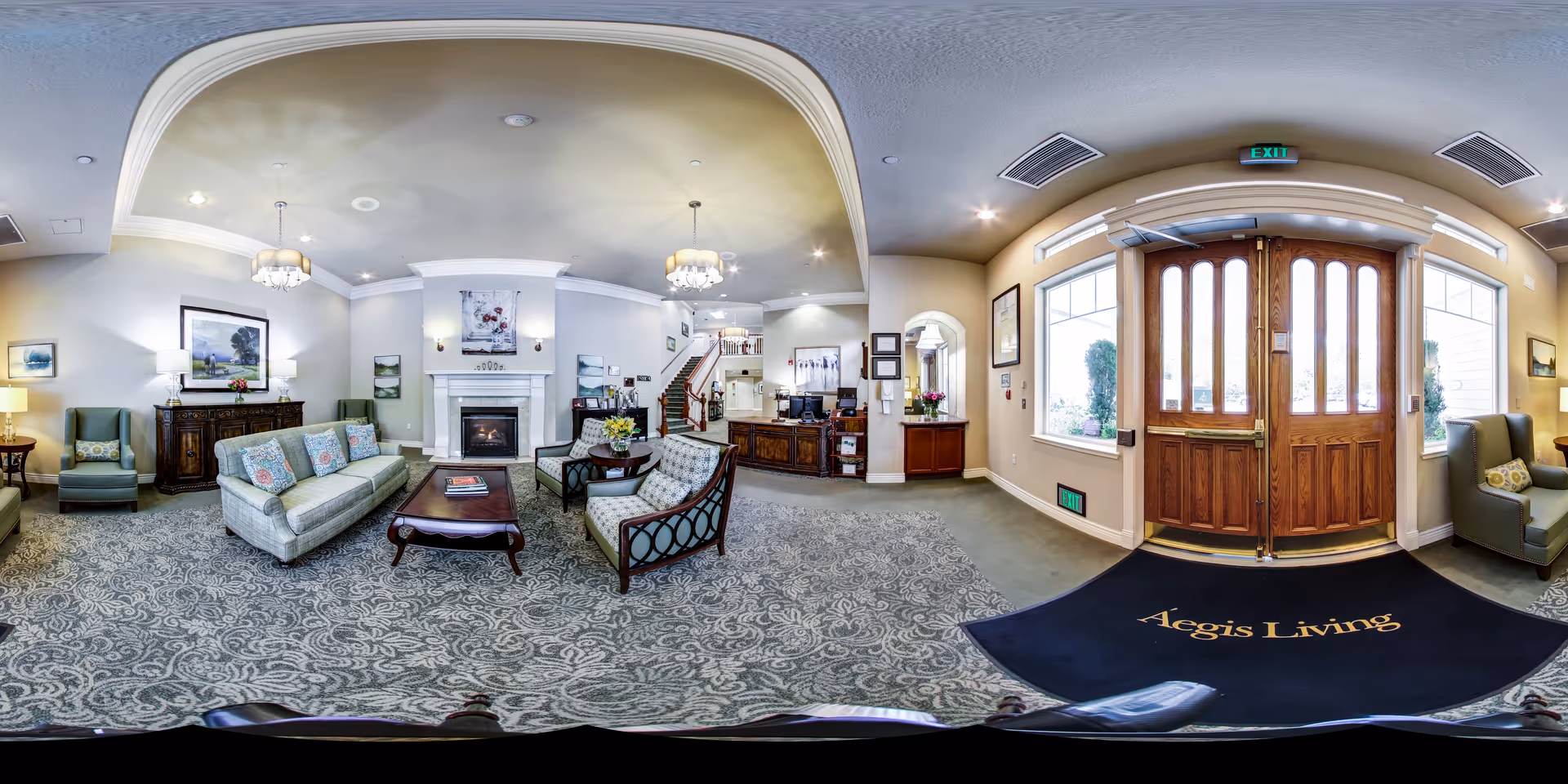 A spacious and elegantly decorated senior living facility lobby with a patterned carpet, comfortable seating including sofas and armchairs, a wooden coffee table, a fireplace with artwork above it, and a reception desk. Large wooden double doors with glass panels allow natural light to enter, and a dark blue mat with the words 'Aegis Living' is placed at the entrance. The room features soft lighting from ceiling fixtures and table lamps, with framed artwork on the walls.