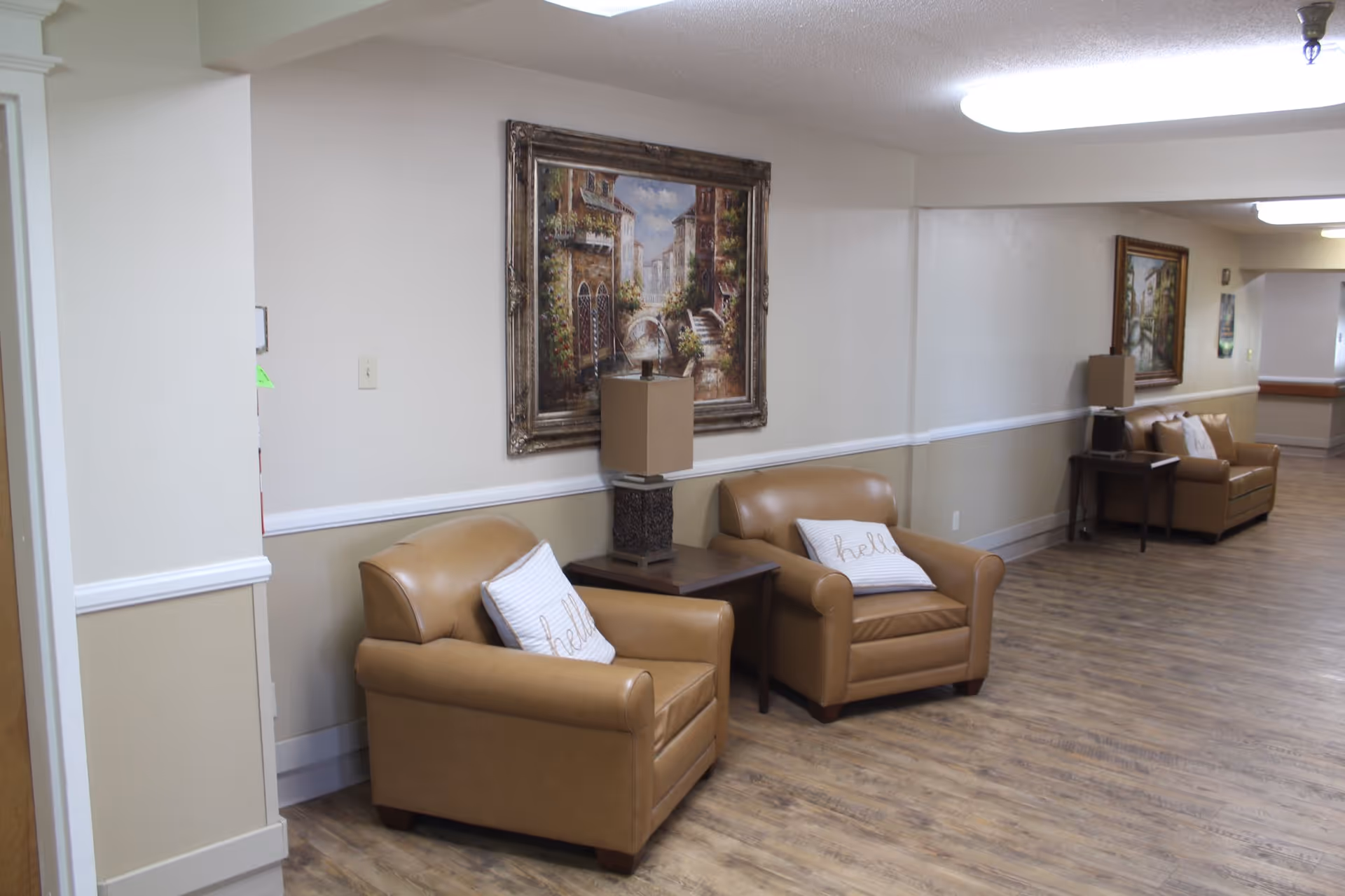 Hallway seating area with tan leather armchairs, side tables, framed paintings, and wood-look flooring in a senior living facility.