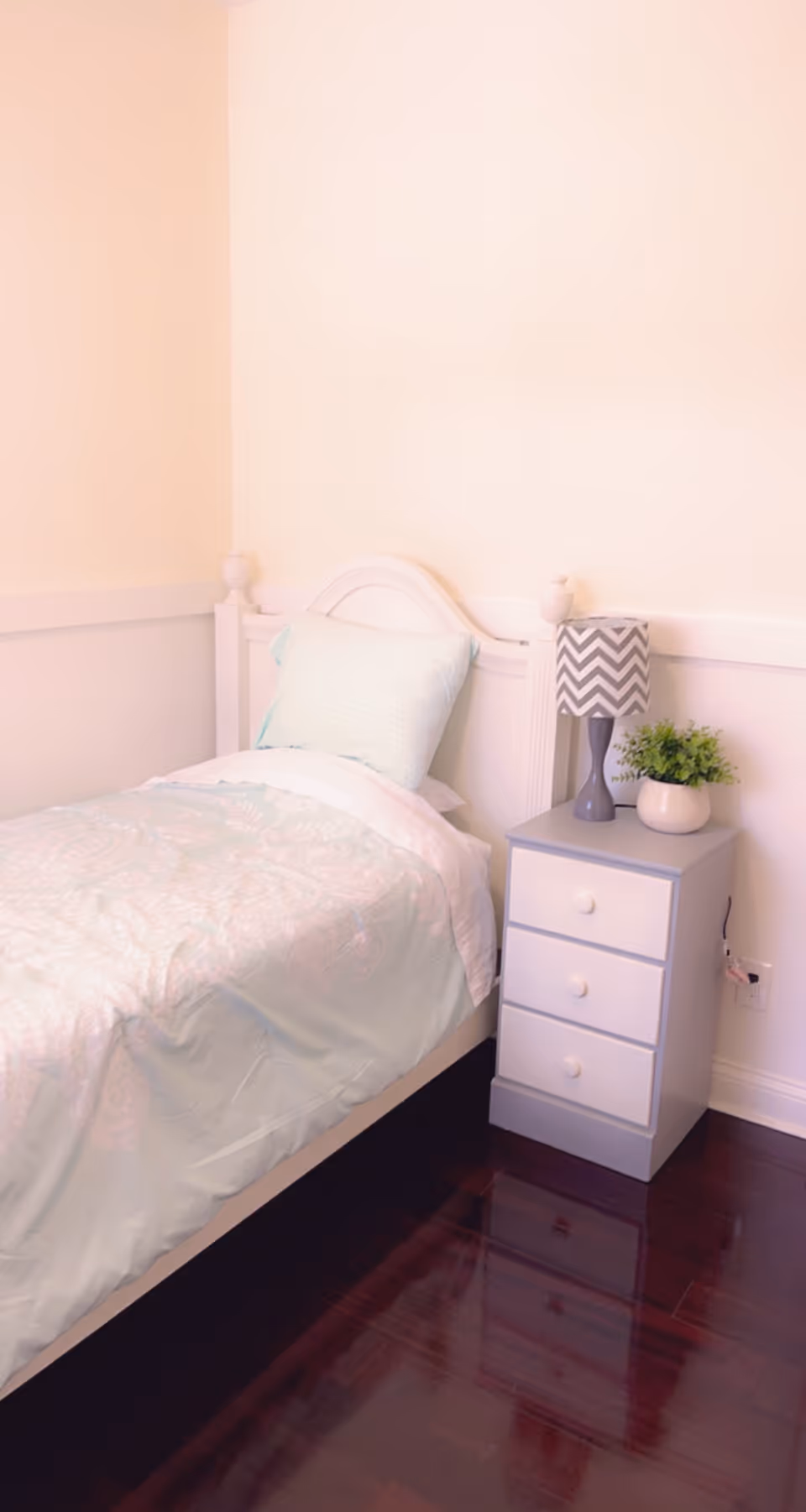 A small bedroom with a single bed featuring a light blue patterned comforter and a matching pillow. Next to the bed is a gray nightstand with three drawers, a gray and white chevron-patterned lamp, and a small potted plant. The room has light-colored walls and dark wooden flooring.