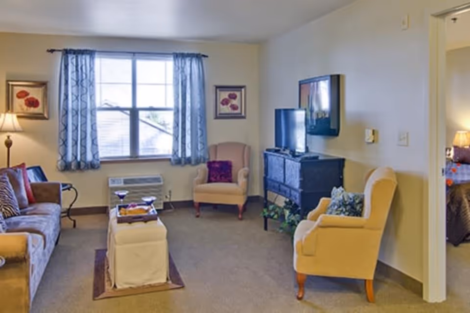 A cozy living room with a sofa, two armchairs, a small ottoman with a tray holding two glasses, a window with patterned curtains, a floor lamp, framed floral artwork on the walls, and a TV mounted above a black cabinet. A glimpse of a bedroom with a bed and lamp is visible through an open doorway.