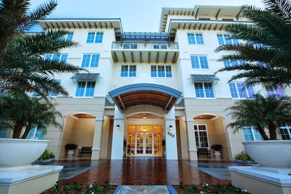 Entrance of a multi-story senior living building with a covered porte-cochère, palm trees, and large planters.