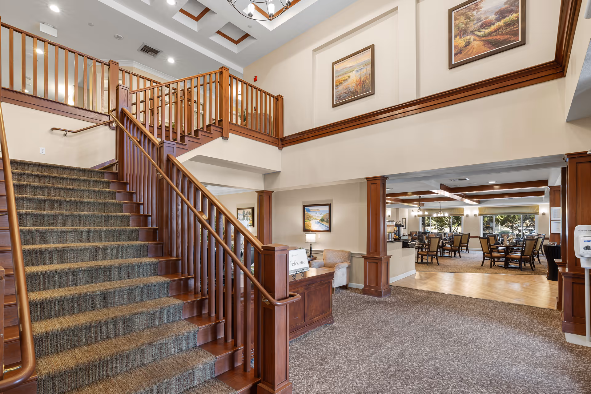 Interior view of a senior living facility featuring a carpeted staircase with wooden railings leading to an upper floor. The area includes a small seating nook with a beige armchair and a wooden welcome desk. The space is decorated with framed landscape paintings and has a chandelier hanging from the ceiling. In the background, there is a dining area with tables and chairs, large windows, and ceiling beams.
