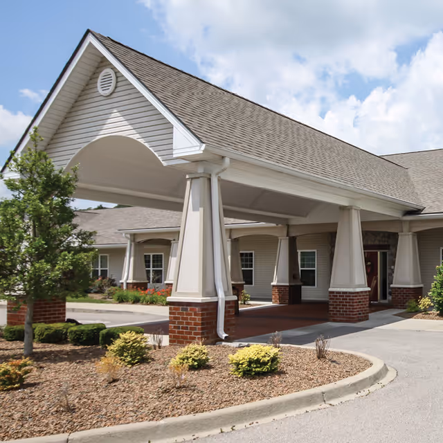 Covered entrance of a senior living facility with large white columns on brick bases, a driveway, landscaped bushes, and a partly cloudy sky.