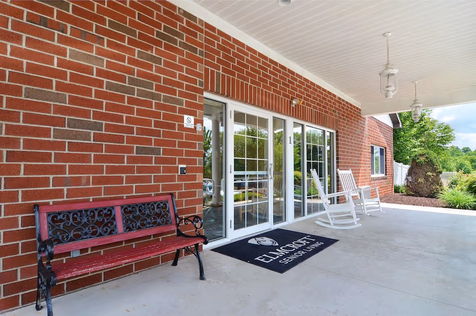 Covered outdoor entrance area of a senior living facility with a red brick wall, a decorative metal and wood bench, two white rocking chairs, glass sliding doors, and a black doormat with the text 'ELMCROFT SENIOR LIVING'. There are hanging lantern-style lights on the white ceiling and greenery visible in the background.