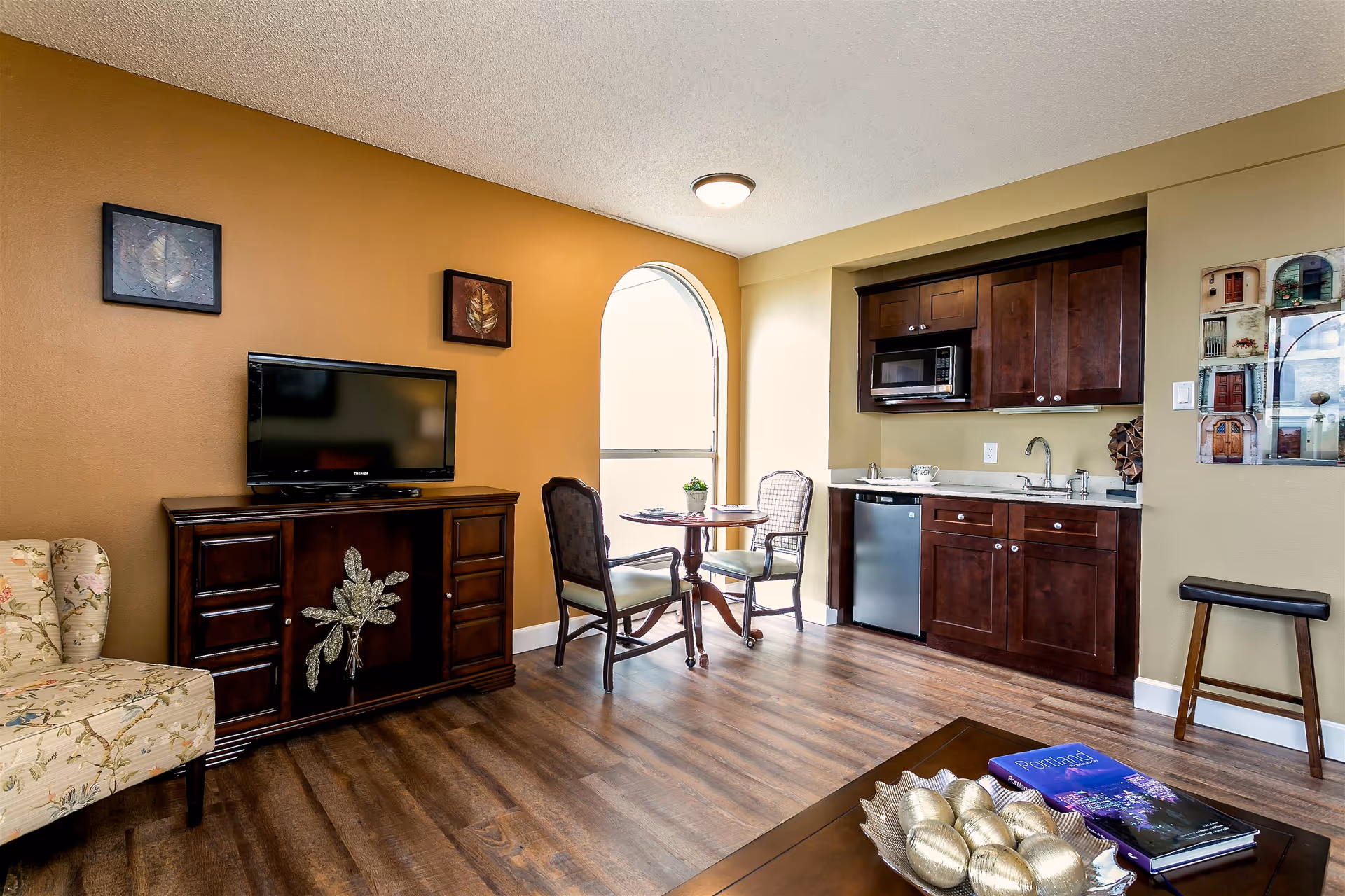 A cozy living area with a wooden floor, featuring a floral upholstered armchair, a dark wood TV stand with a flat-screen TV, two chairs and a small round table near an arched window, and a kitchenette with dark wood cabinets, a microwave, a small refrigerator, and a sink. The walls are painted in warm tones with decorative artwork.