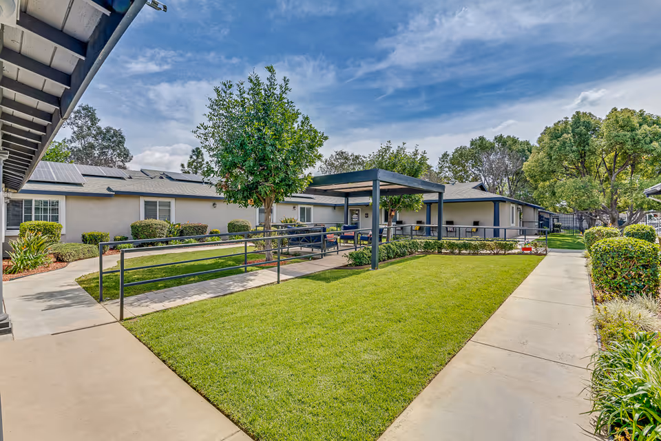 Outdoor courtyard area at a senior living facility with well-maintained green grass, a paved walkway, a covered seating area with benches, and surrounding single-story buildings with windows and solar panels on the roof. Trees and shrubs are planted around the courtyard under a partly cloudy blue sky.