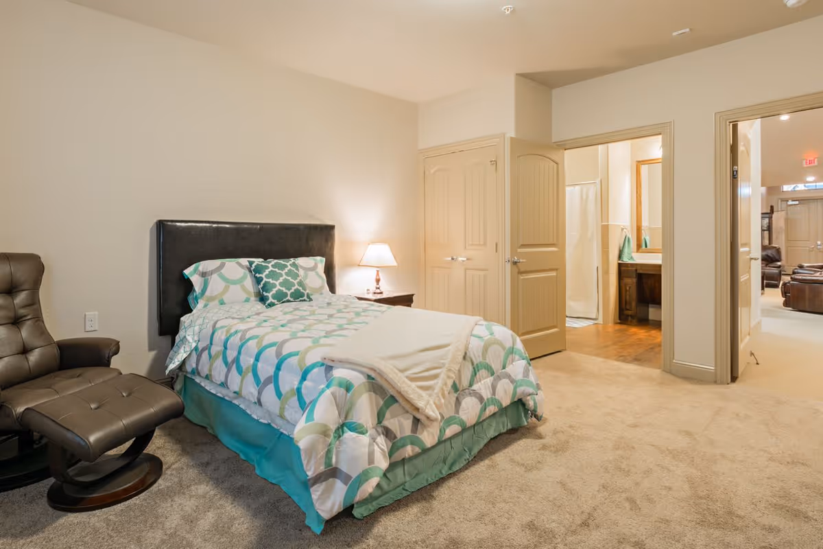 A bedroom with a single bed featuring a patterned comforter and pillows, a bedside table with a lamp, a brown leather recliner chair, carpeted floor, and two open doors leading to a bathroom and another room with seating.
