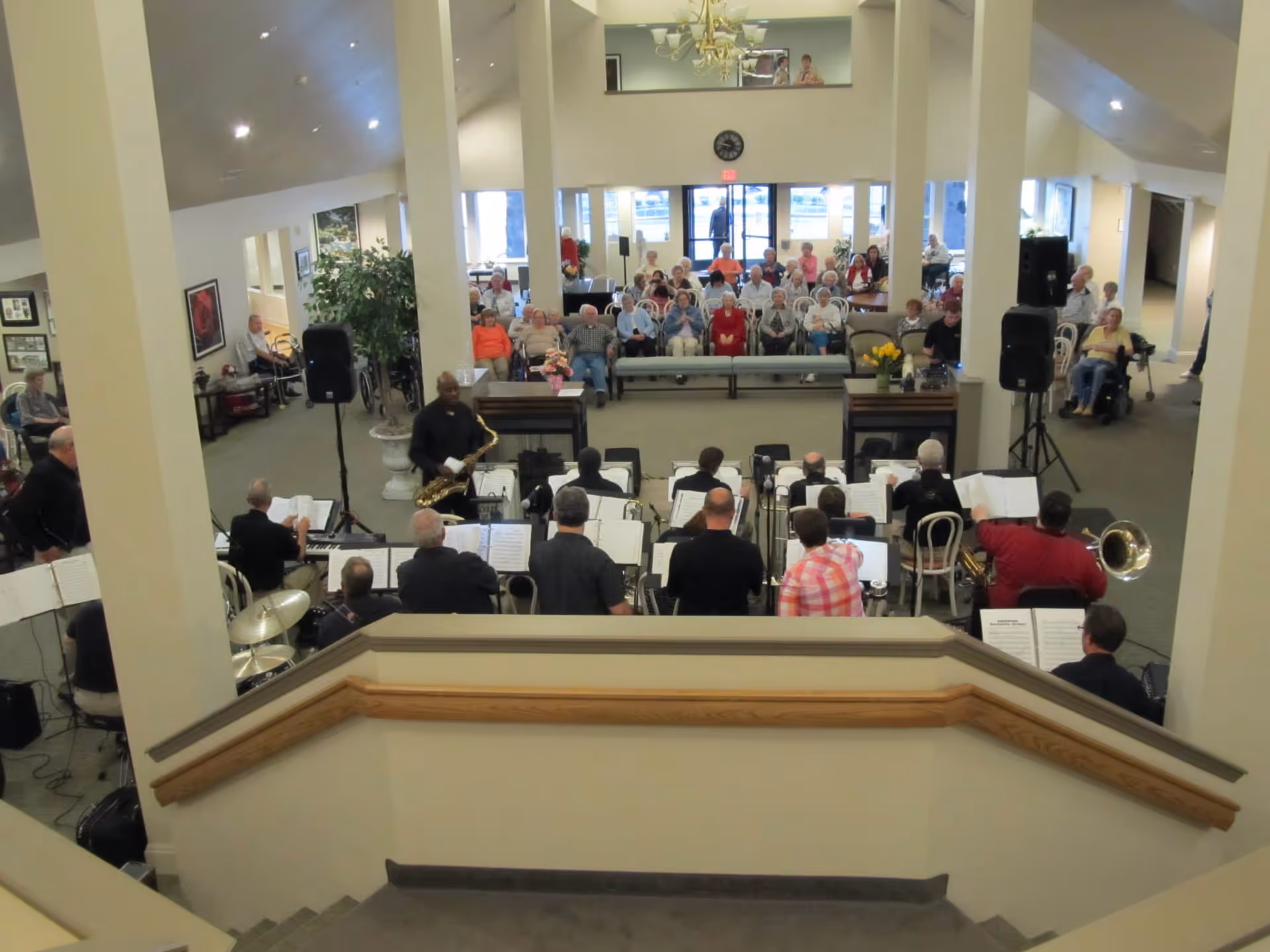 A musical performance in a senior living facility common area with a band playing instruments such as saxophone, drums, and keyboard. The audience, consisting mostly of elderly people, is seated facing the band. The room has high ceilings, large windows, and light-colored walls with columns.
