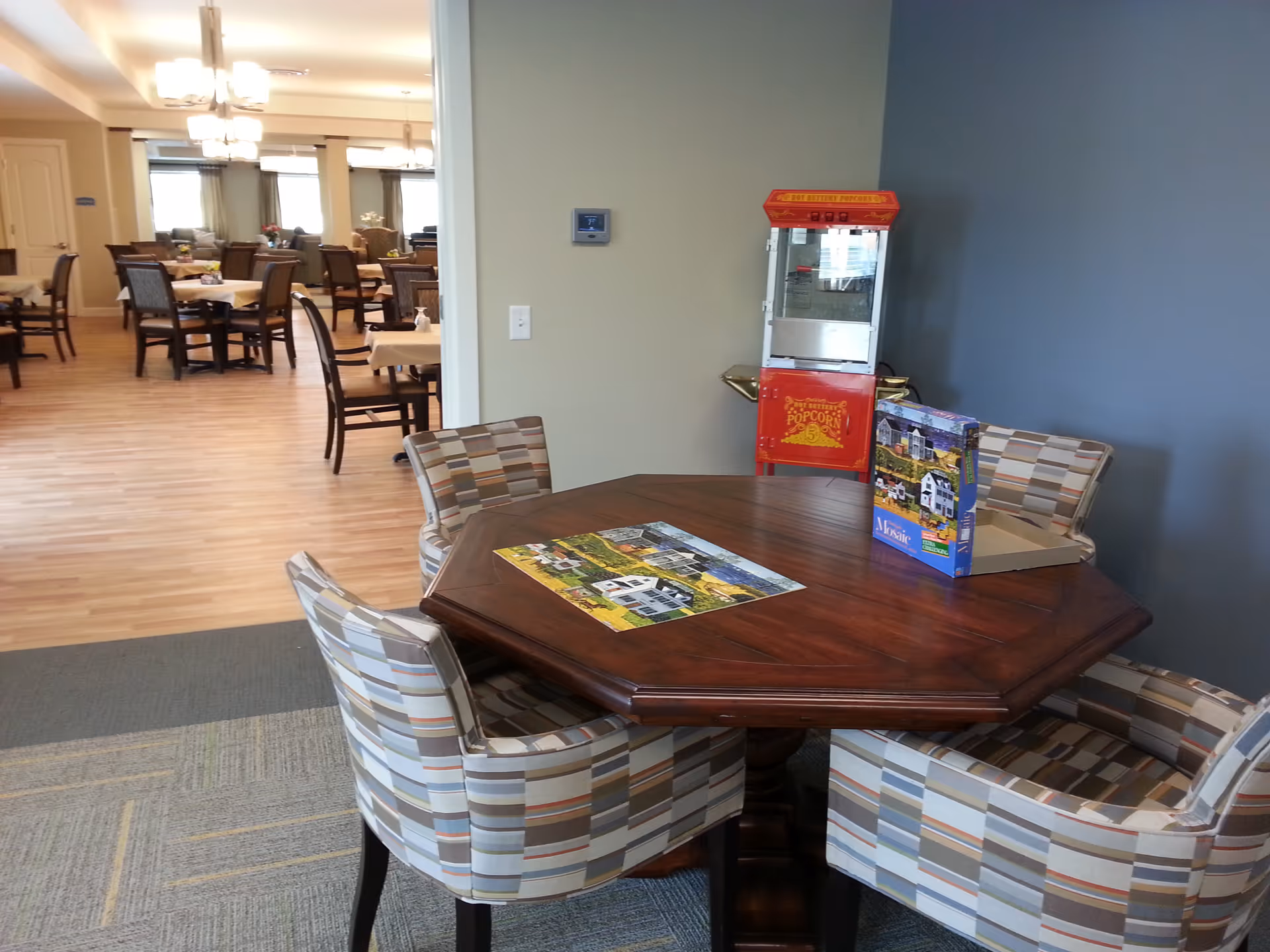 A cozy common area in Emery Place featuring a wooden octagonal table surrounded by four patterned upholstered chairs. On the table, there is a partially completed puzzle and its box. In the background, there is a red popcorn machine against a blue-gray wall. Beyond this area, a dining room with multiple tables and chairs is visible, illuminated by ceiling lights.