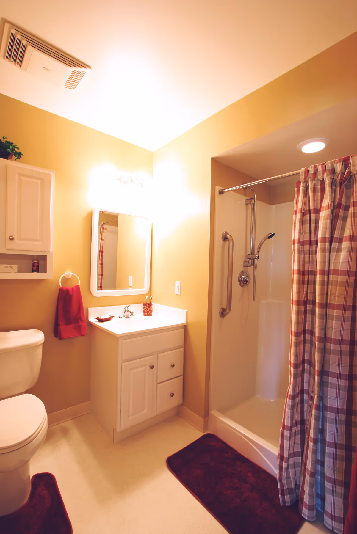 A small bathroom with beige walls featuring a white toilet, a white vanity with a sink and mirror above it, a shower with a plaid shower curtain, and red bath mats on the floor. A red hand towel hangs on a ring next to the vanity, and there is a white cabinet mounted on the wall above the toilet.