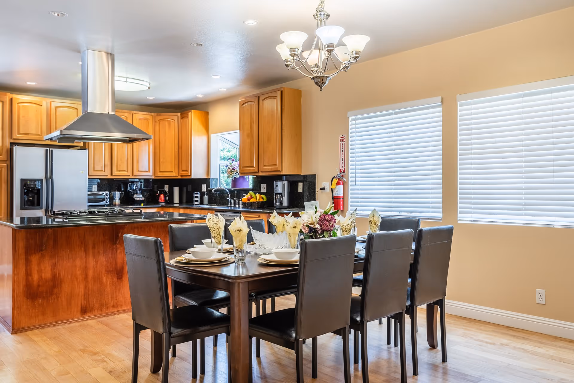 A bright and spacious kitchen and dining area with wooden cabinets, a stainless steel refrigerator, and a kitchen island with a stove and range hood. The dining table is set with plates, bowls, glasses with folded napkins, and a floral centerpiece. Two large windows with white blinds allow natural light into the room.