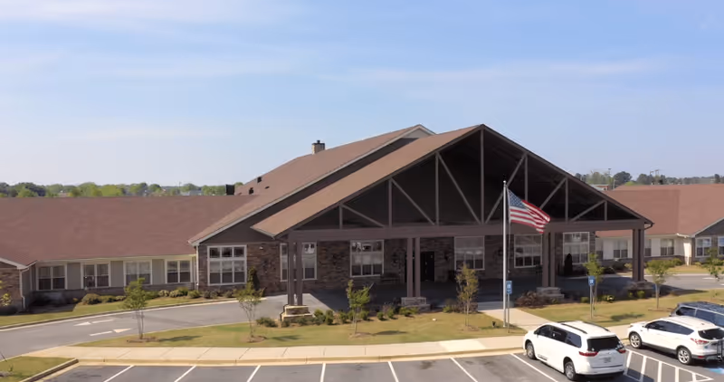 Front exterior view of a single-story senior living facility building with a large covered entrance, an American flag on a flagpole, landscaped greenery, and a parking lot with several cars.