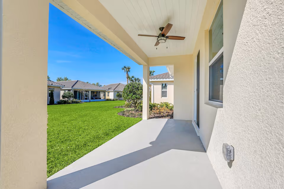 Covered outdoor patio area with a ceiling fan, overlooking a well-maintained green lawn and neighboring single-story buildings under a clear blue sky.
