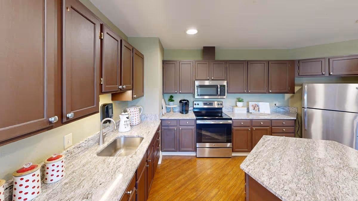 A modern kitchen with brown cabinets, granite countertops, and stainless steel appliances including a refrigerator, stove, and microwave. The kitchen features a double sink, wooden flooring, and various decorative items on the counters.