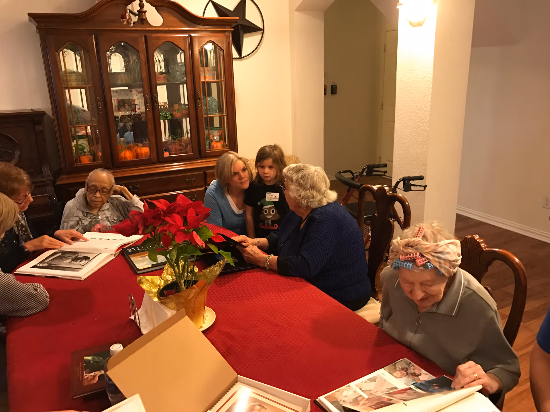A group of elderly women and a young girl gathered around a dining table covered with a red tablecloth, looking at photo albums. The room has wooden furniture including a china cabinet filled with decorative items and a poinsettia plant centerpiece on the table.