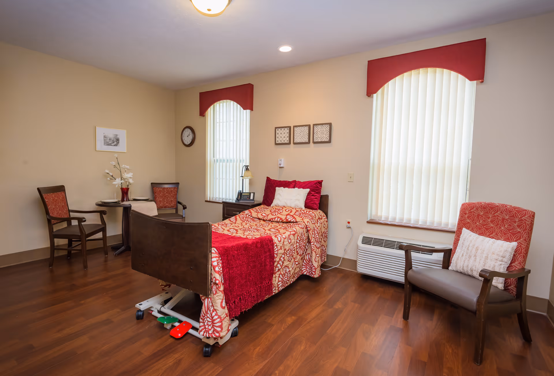 A senior living facility bedroom with a single bed covered in a red and beige patterned bedspread and red throw. The room has two windows with vertical blinds and red valances. There are two chairs with red cushions, a small round table with a vase of flowers, a clock on the wall, and a heating/cooling unit under one window. The floor is wood, and the walls are beige.