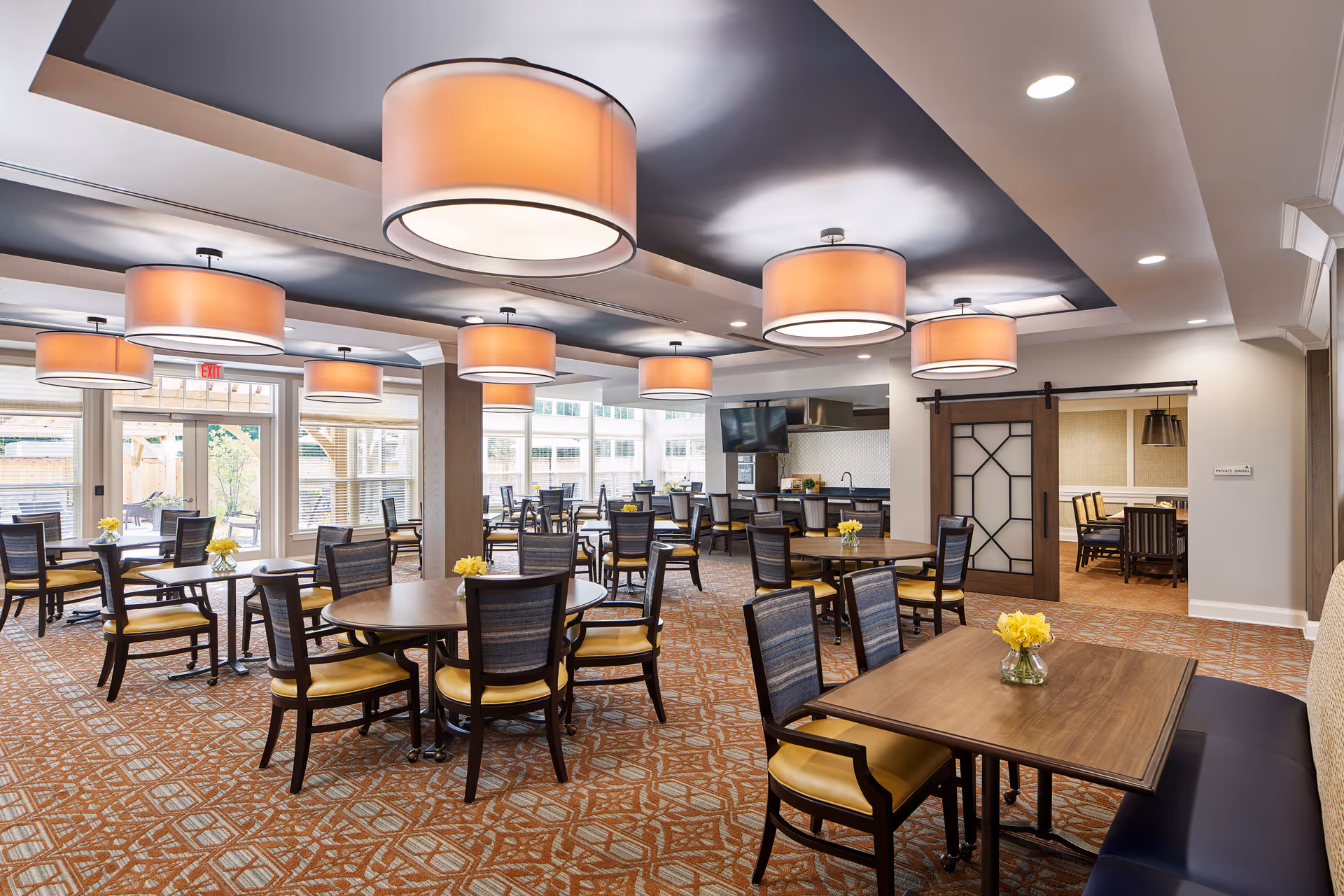 A spacious dining room with multiple round and square wooden tables surrounded by chairs with yellow cushions. The room features large, round ceiling lights with warm lighting, a patterned carpet, and large windows letting in natural light. There are small vases with yellow flowers on each table. A sliding barn door leads to another room with additional seating.
