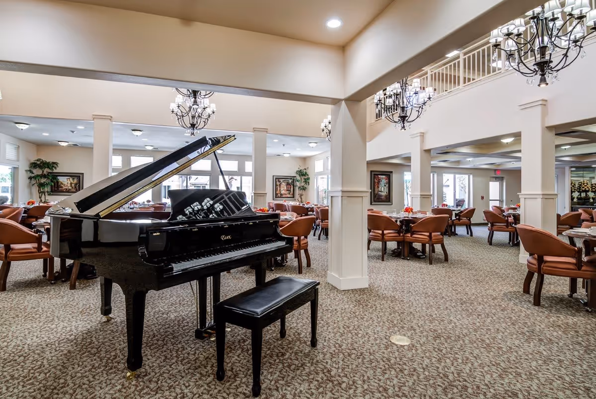 Spacious senior living facility common area with a black grand piano and matching bench in the foreground. The room features multiple round tables with brown cushioned chairs arranged around them, beige carpet, white pillars, and elegant chandeliers hanging from the ceiling. Large windows allow natural light to brighten the space, and framed artwork and plants decorate the walls.